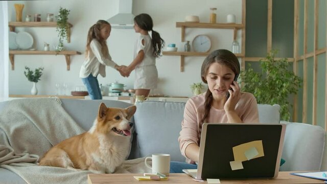 A Busy Mother Trying to Work Remotely at Home. On The Background Her Daughters are Jumping Together, While Mother Working With a Laptop and Doing Business Conversation on the Phone