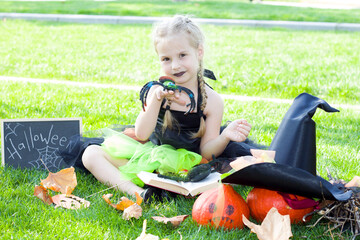 little girl in a witch costume in the Park on the lawn. In the hands of holds a spider. Next to it is a broom, a hat, and pumpkins. Halloween