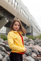 Pretty young sexy woman of athletic build is standing on stones on city beach. Walk through city center. Portrait of cute female in yellow jacket and black trousers on street