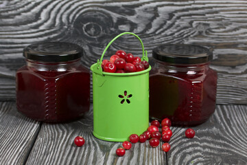 Red cranberries in a miniature green bucket. Stands on brushed boards. Nearby is jars of cranberry jam and some berries scattered around.