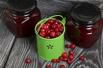 Red cranberries in a miniature green bucket. Stands on brushed boards. Nearby is jars of cranberry jam and some berries scattered around.