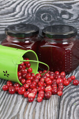 Red cranberries in an overturned green bucket. Lies on brushed boards. Next to it is cranberry jam in a jar.