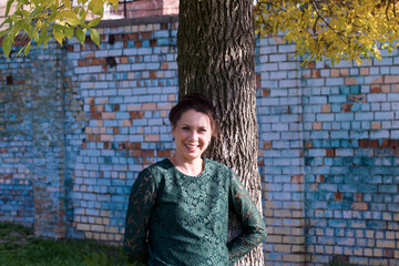 Fototapeta na wymiar The girl in a green tracksuit. Stands near a tree with yellowed foliage. Against the background of a brick wall.