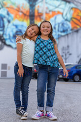 Obraz na płótnie Canvas Two young sisters hold each other side by side in a colorful outdoor parking lot. Standing together with arms wrap around each other, looking at the camera with joy.