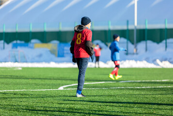 Boys in red sportswear running on soccer field with snow on background. Young footballers dribble and kick football ball in game. Training, active lifestyle, sport, children winter activity © Natali