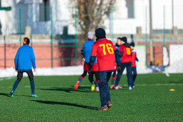 Boys in red sportswear running on soccer field with snow on background. Young footballers dribble and kick football ball in game. Training, active lifestyle, sport, children winter activity