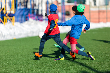 Boys in red sportswear running on soccer field with snow on background. Young footballers dribble...