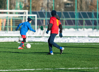Obraz premium Boys in red sportswear running on soccer field with snow on background. Young footballers dribble and kick football ball in game. Training, active lifestyle, sport, children winter activity