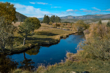 Fototapeta na wymiar Bright blue winding river flows in grass, bushes, trees among the mountains and sky with clouds. Reflections in clear water. Autumn russia landscape