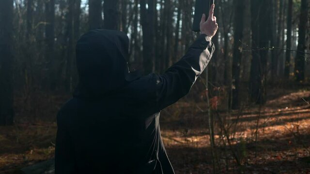 Monk in Black Cowl Hood Rises up Hand Holding Bible. Sunshine in Misty Wood. Autumn Morning. 2x Slow motion 60 fps