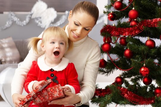 Happy little girl holding christmas surprise sitting by christmas tree with mother.