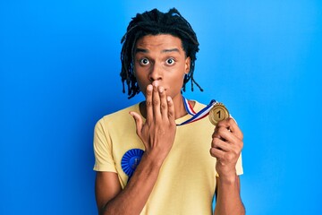 Young african american man wearing first place badge holding medal covering mouth with hand, shocked and afraid for mistake. surprised expression