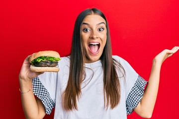 Young hispanic woman eating hamburger celebrating victory with happy smile and winner expression with raised hands