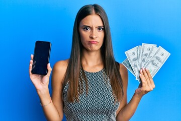 Young hispanic woman holding dollars and using smarpthone skeptic and nervous, frowning upset because of problem. negative person.
