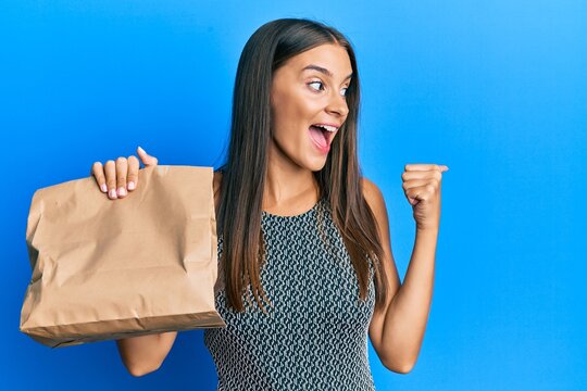 Young hispanic woman holding take away paper bag pointing thumb up to the side smiling happy with open mouth