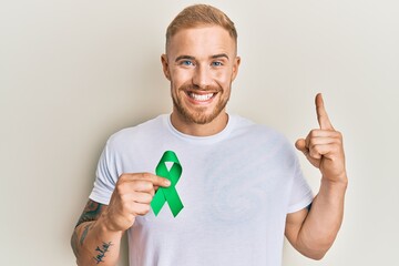 Young caucasian man holding support green ribbon smiling with an idea or question pointing finger...