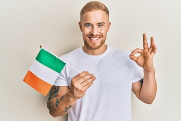 Young caucasian man holding irish flag doing ok sign with fingers, smiling friendly gesturing excellent symbol