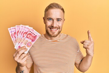 Young caucasian man holding 100 yuan chinese banknotes smiling with an idea or question pointing finger with happy face, number one