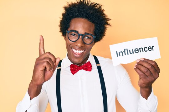 Handsome african american man with afro hair holding paper with influencer text smiling with an idea or question pointing finger with happy face, number one