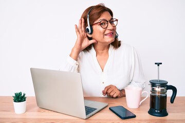 Middle age brunette hispanic woman working at the office wearing operator headset smiling with hand over ear listening and hearing to rumor or gossip. deafness concept.