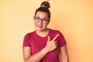 Middle age brunette hispanic woman wearing casual clothes and glasses smiling cheerful pointing with hand and finger up to the side