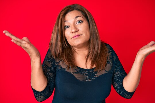 Middle age brunette hispanic woman wearing elegant clothes over red background clueless and confused with open arms, no idea and doubtful face.