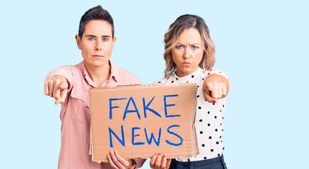 Couple of women holding fake news banner pointing with finger to the camera and to you, confident gesture looking serious