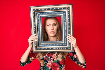 Fototapeta premium Young caucasian woman with blond hair holding empty frame skeptic and nervous, frowning upset because of problem. negative person.