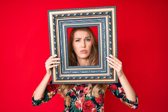 Young caucasian woman with blond hair holding empty frame skeptic and nervous, frowning upset because of problem. negative person.