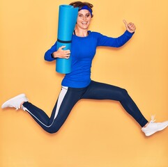Young beautiful sportswoman smiling happy. Jumping with smile on face holding yoga mat doing ok sign with thumbs up over isolated yellow background.