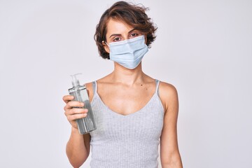Young hispanic woman wearing medical mask holding hand sanitizer gel thinking attitude and sober expression looking self confident