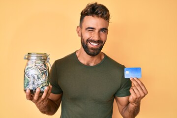 Young hispanic man holding credit car and jar with dollars winking looking at the camera with sexy...