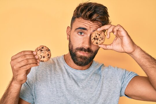 Young hispanic man holding cookie clueless and confused expression. doubt concept.