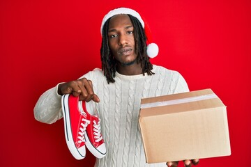 African american man with braids wearing christmas hat having shoes as a gift relaxed with serious expression on face. simple and natural looking at the camera. - obrazy, fototapety, plakaty