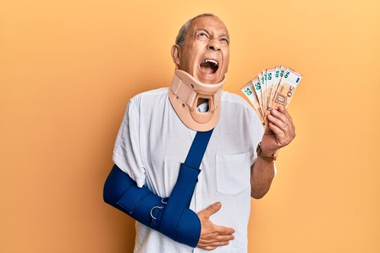 Handsome mature injured man wearing cervical collar and sling holding insurance euros angry and mad screaming frustrated and furious, shouting with anger looking up.