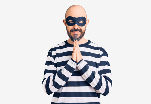 Young handsome man wearing burglar mask praying with hands together asking for forgiveness smiling confident.