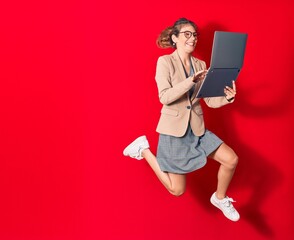 Young beautiful blonde businesswoman wearing elegant clothes and glasses smiling happy. Jumping with smile on face using laptop over isolated red background