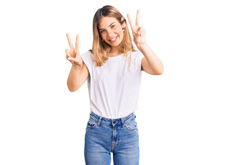 Beautiful caucasian woman with blonde hair wearing casual white tshirt smiling looking to the camera showing fingers doing victory sign. number two.