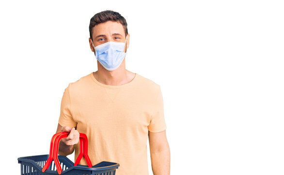 Handsome young man with bear wearing shopping basket and medical mask looking positive and happy standing and smiling with a confident smile showing teeth