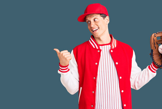 Young handsome man wearing baseball uniform holding golve and ball pointing thumb up to the side smiling happy with open mouth