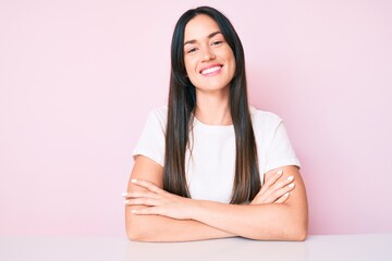 Young caucasian woman sitting at the table wearing casual white tshirt happy face smiling with crossed arms looking at the camera. positive person.