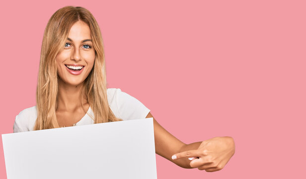 Beautiful blonde young woman holding blank empty banner smiling happy pointing with hand and finger