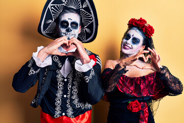 Young couple wearing mexican day of the dead costume over yellow smiling in love doing heart symbol shape with hands. romantic concept.