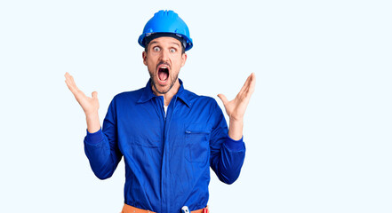 Young handsome man wearing worker uniform and hardhat celebrating crazy and amazed for success with arms raised and open eyes screaming excited. winner concept