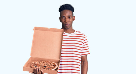 Young african american man holding delivery pizza box thinking attitude and sober expression looking self confident