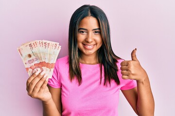 Young latin girl holding 10 colombian pesos banknotes smiling happy and positive, thumb up doing excellent and approval sign