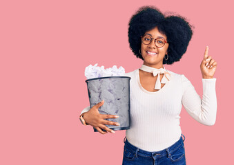 Young african american girl holding paper bin full of crumpled papers surprised with an idea or question pointing finger with happy face, number one