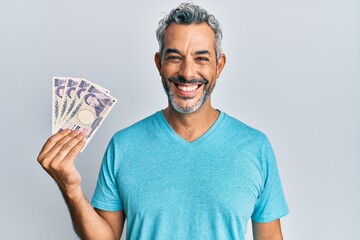 Middle age grey-haired man holding japanese yen banknotes looking positive and happy standing and smiling with a confident smile showing teeth