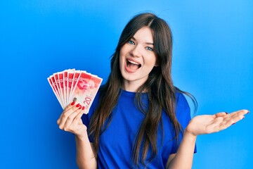 Young beautiful caucasian girl holding 20 israel shekels banknotes celebrating achievement with happy smile and winner expression with raised hand
