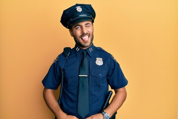 Handsome hispanic man wearing police uniform sticking tongue out happy with funny expression. emotion concept.